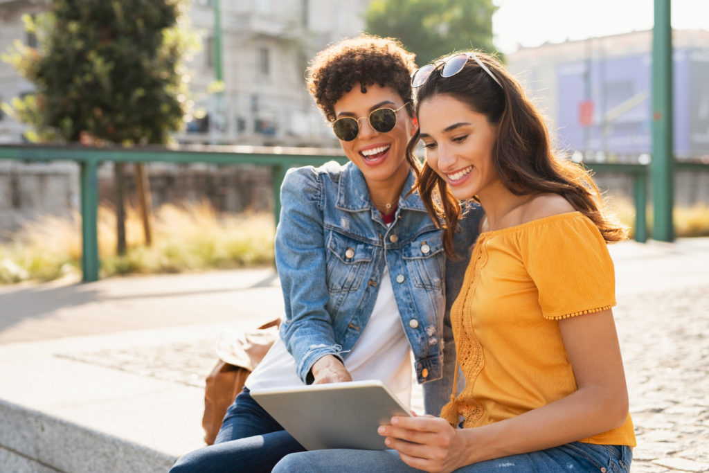 Two happy girls using digital tablet while sitting on stairs outdoor. Latin young woman looking for new sales on online store with her african friend. Multiethnic teens having fun while using laptop sitting on city street at sunset.