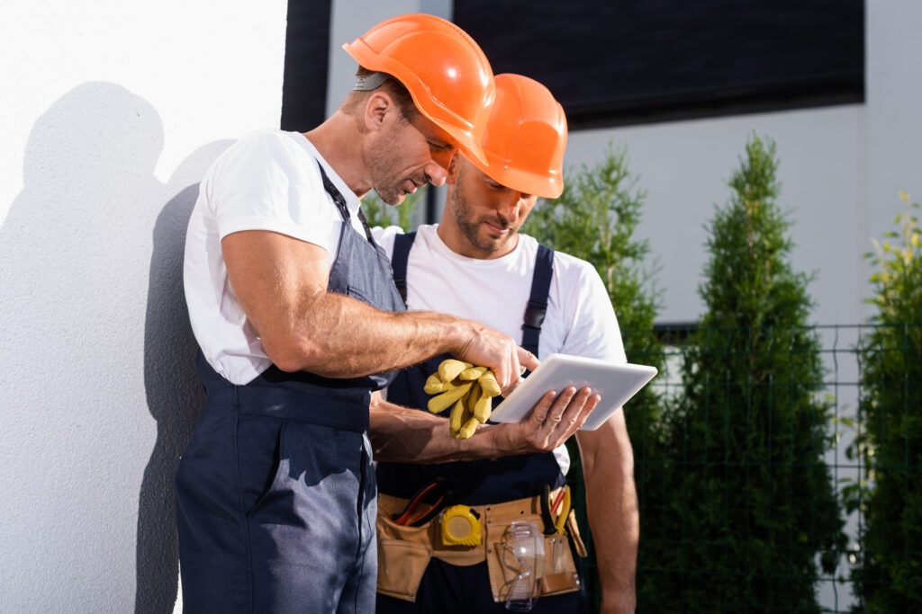 handyman in overalls using digital tablet while standing near facade of house