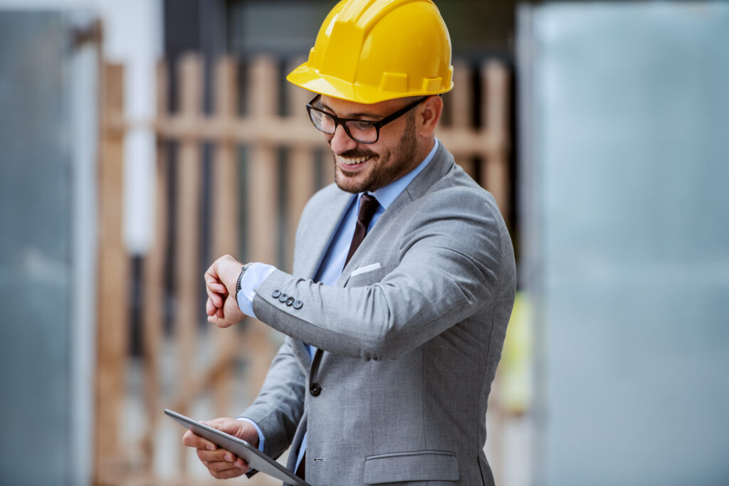 attractive elegant caucasian smiling architect in suit with eyeglasses and helmet on head standing at construction site and holding tablet while looking at wristwatch