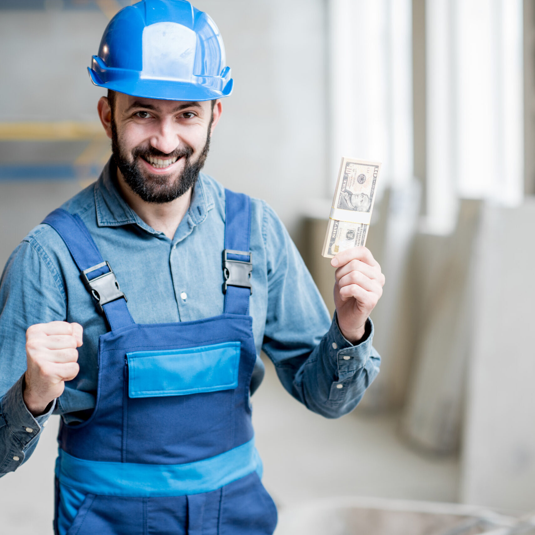 Builder with salary at the construction site Happy construction worker in blue overalls standing with his salary at the construction site indoors